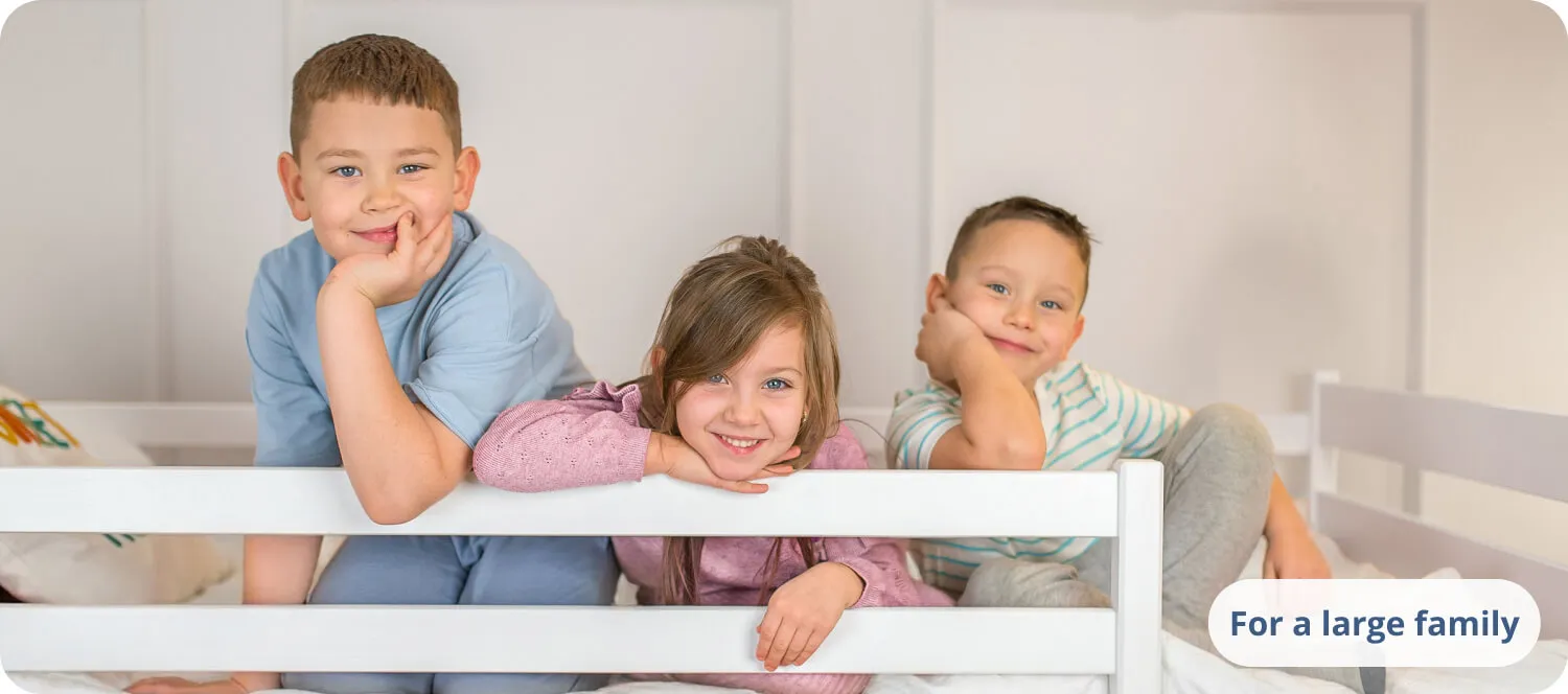Three smiling children leaning on a white bed railing.